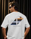 A man wearing a white t-shirt from the back, featuring a large graphic print with a butterfly, a split mountain design, and the text 'THE AVICII TRIBUTE' against a dark curtain background.
