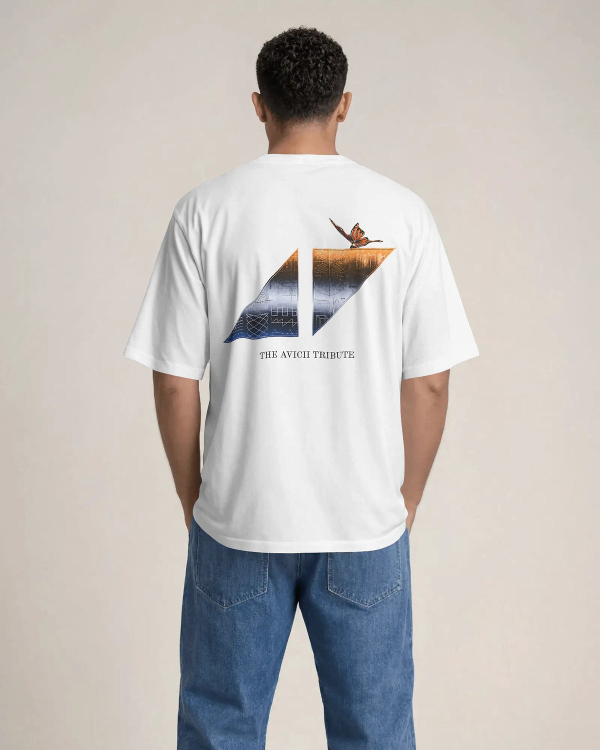 A man viewed from behind wearing a white t-shirt with a large graphic on the back and blue jeans, standing against a plain beige studio background. The Avicii Tribute Tomorrowland Tshirt.