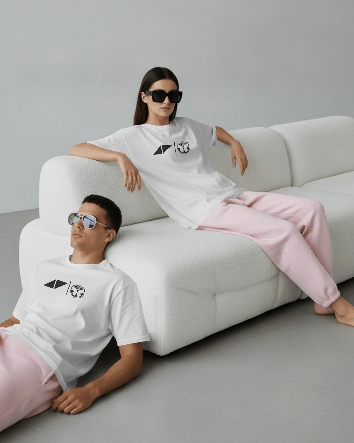 A man and a woman wearing matching white graphic t-shirts and light pink sweatpants, posed on a modern white textured sofa in a minimalist gray room.