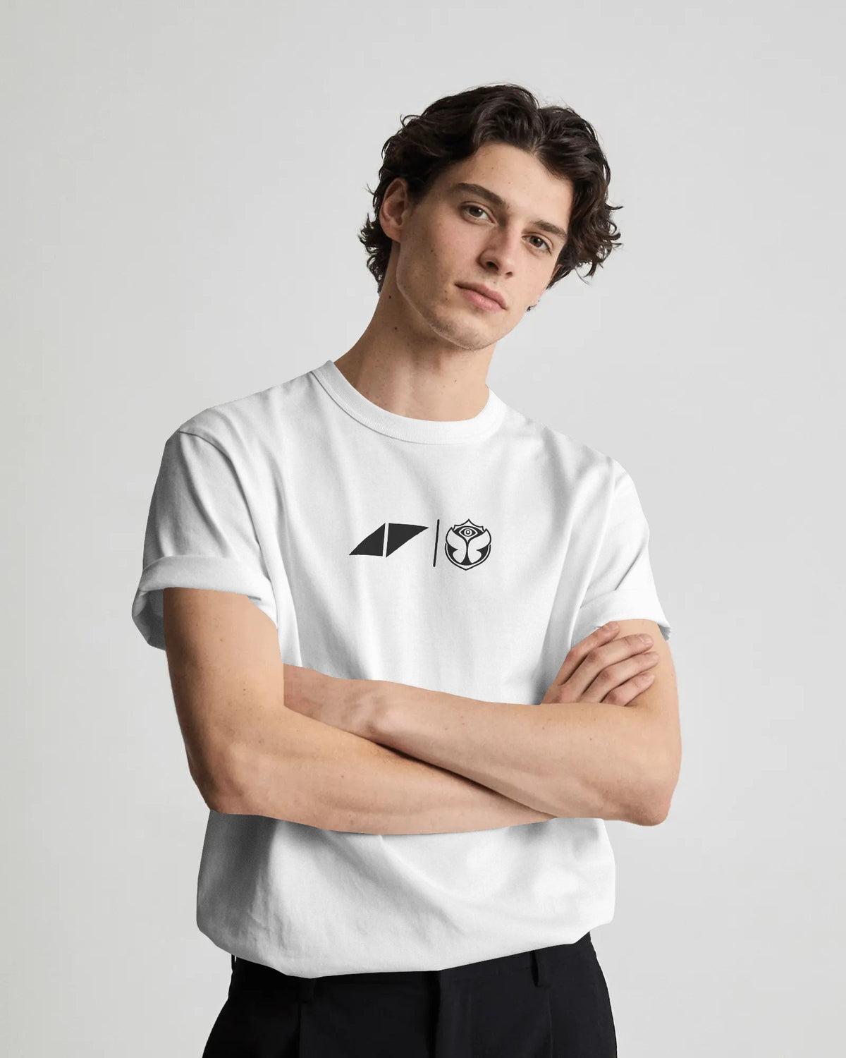 A man wearing a white short-sleeve crewneck t-shirt with a black geometric logo and a wolf head graphic on the chest, paired with black pants, against a light gray studio background.