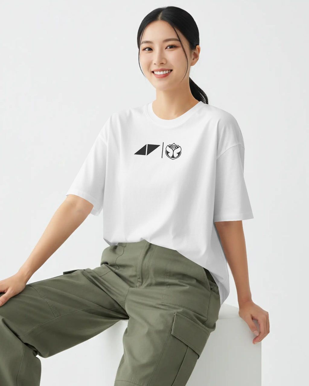 A woman wearing a white short-sleeve t-shirt with a black geometric chest graphic and olive green cargo pants, sitting against a clean white background.