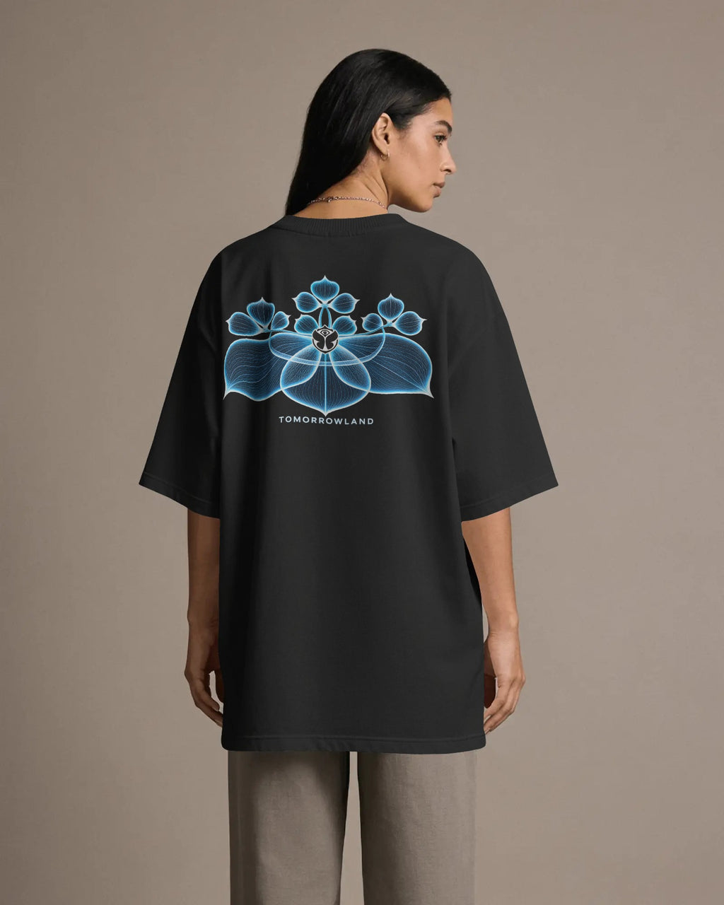 A woman shown from the back wearing an oversized black T-shirt with a large blue floral graphic and the text 'TOMORROWLAND' printed on the back, paired with light gray pants, against a plain beige studio background.