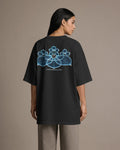 A woman shown from the back wearing an oversized black T-shirt with a large blue floral graphic and the text 'TOMORROWLAND' printed on the back, paired with light gray pants, against a plain beige studio background.