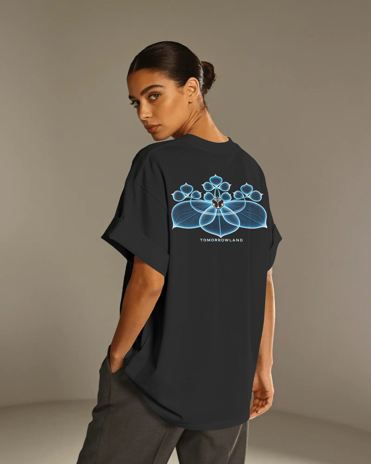 A woman modeling a black oversized T-shirt from the back, featuring a glowing blue geometric mandala graphic and the text 'Tomorrowland' printed below it. She is wearing dark gray pants against a neutral studio backdrop.
