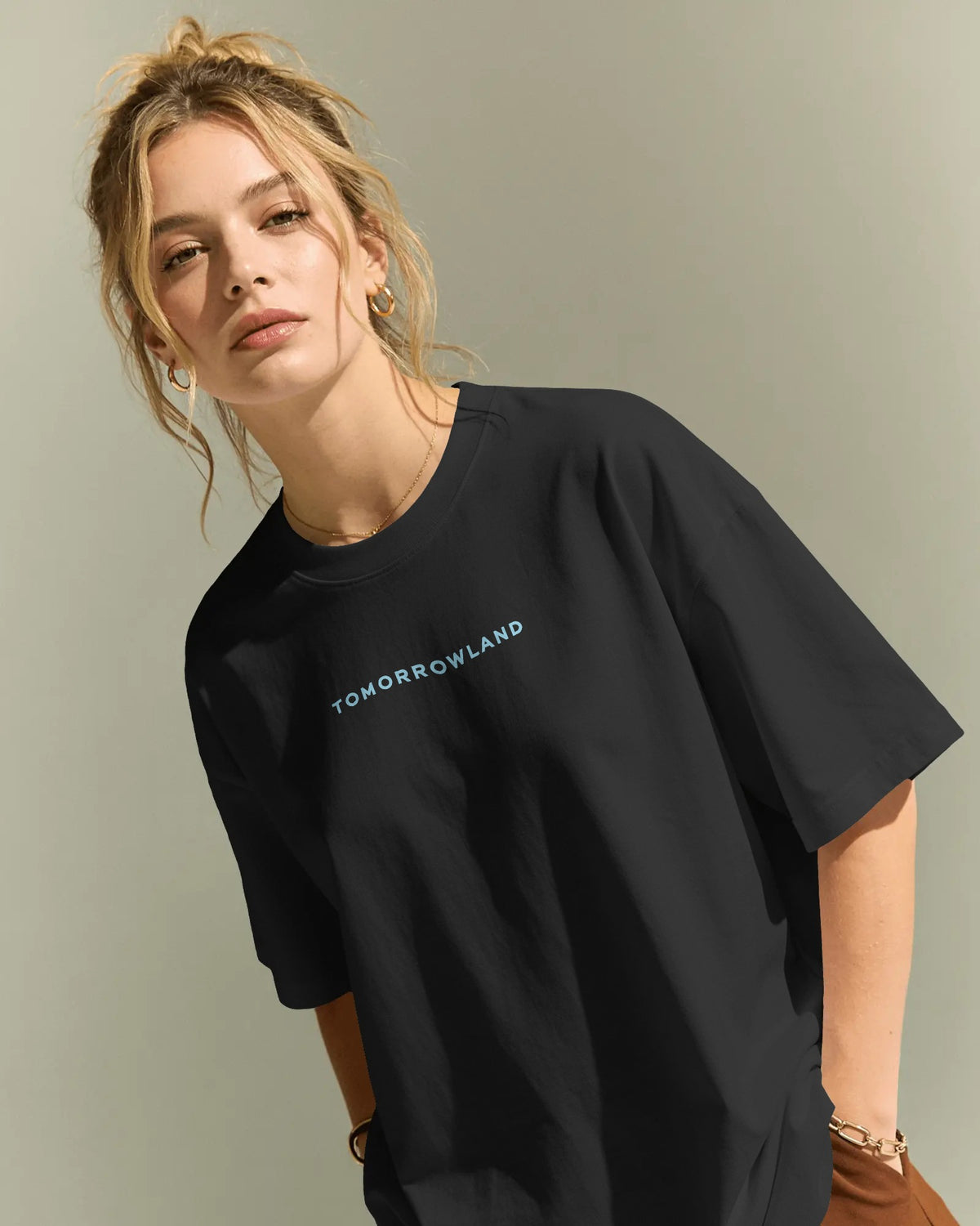 A woman wearing an oversized black t-shirt with 'TOMORROWLAND' printed in light blue across the chest, posed against a neutral gray background. Nitorious Atelier Tomorrowland Concert collection