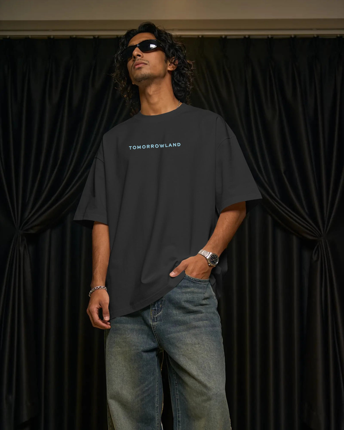 A man wearing an oversized black Tomorrowland t-shirt and blue jeans stands in front of black curtains, with his hand in his pocket.