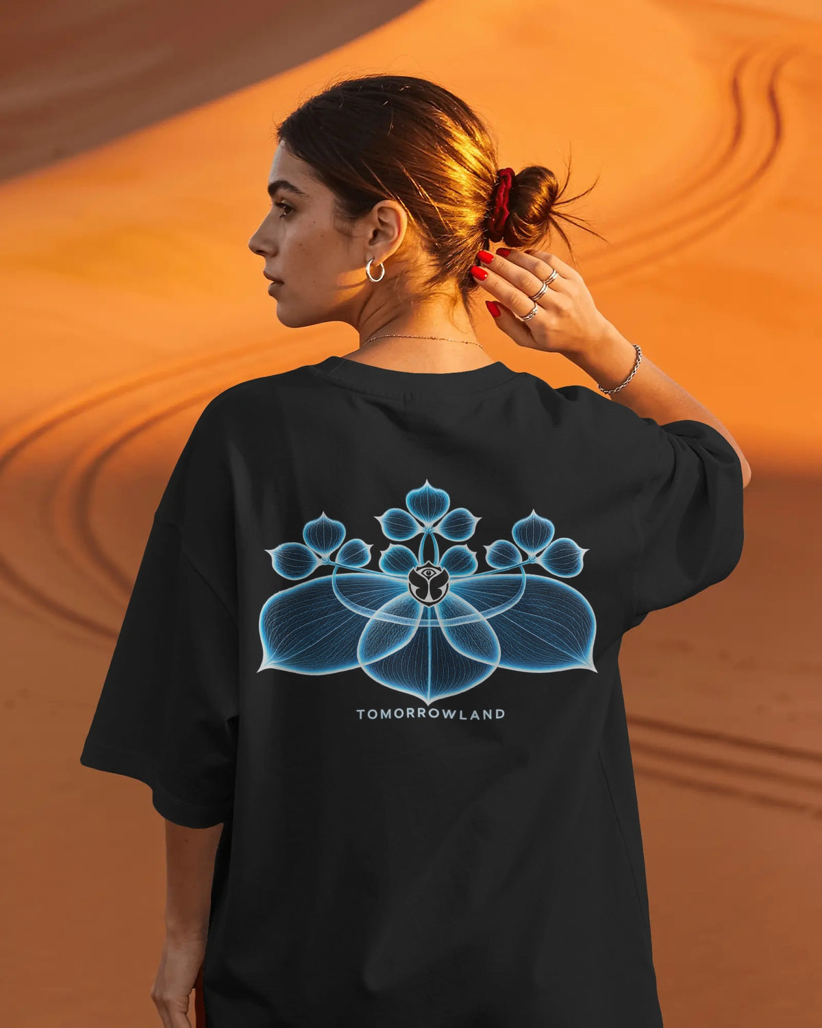 A woman wearing an oversized black Tomorrowland t-shirt, viewed from behind to show the large blue and white geometric flower graphic on the back, set against a warm orange desert dune background.