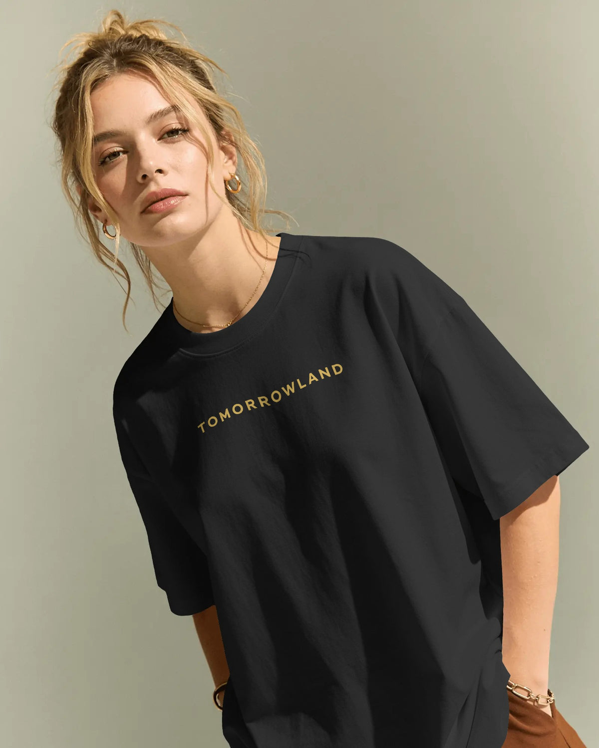 A woman wearing an oversized black t-shirt with 'TOMORROWLAND' printed in gold across the chest, posed against a neutral light gray background.