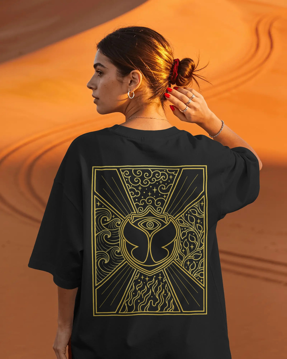 A woman shown from the back wearing an oversized black T-shirt with a large gold occult-style eye graphic printed on the back, set against a warm orange desert dune background. Nitorious Atelier Tomorrowland Concert collection