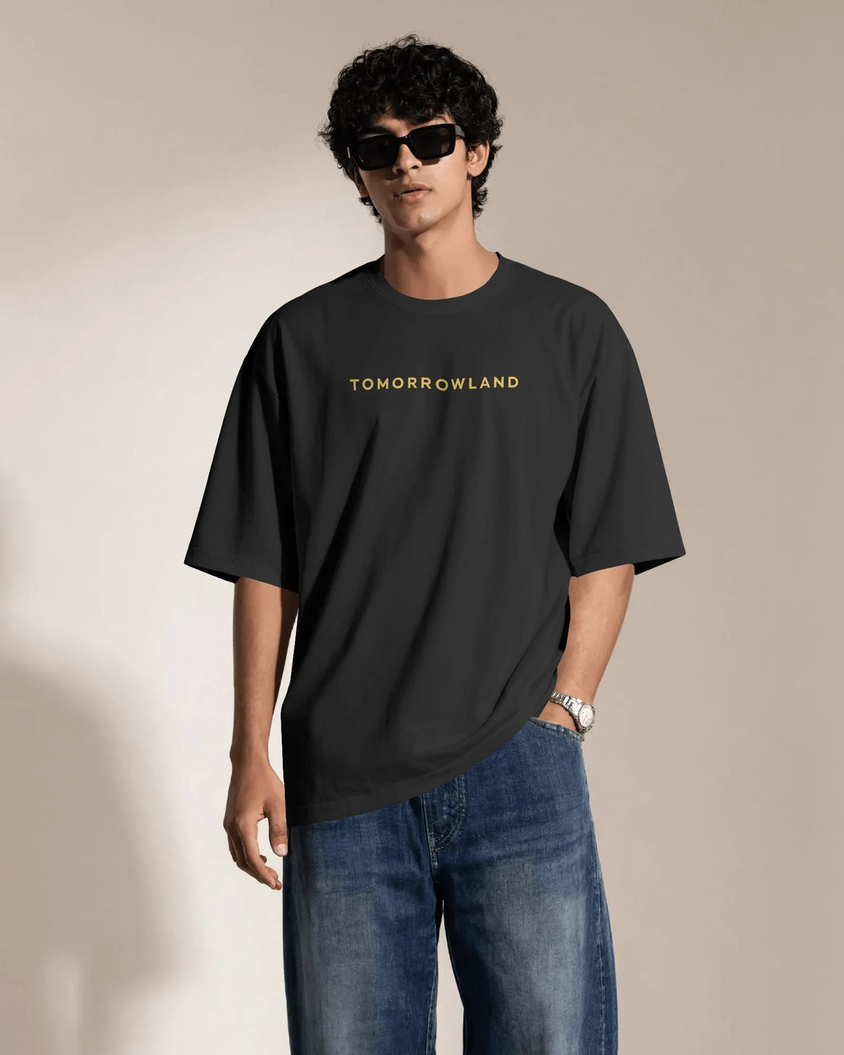 A man wearing a black oversized t-shirt with 'TOMORROWLAND' printed in gold across the chest, paired with blue jeans and black sunglasses, standing against a neutral beige background.