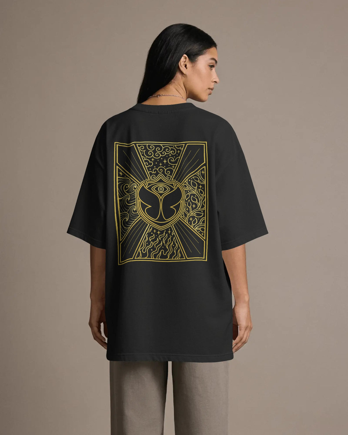 A woman shown from the back wearing an oversized black T-shirt with a large gold graphic print on the back, paired with light gray pants, against a solid beige studio background. Nitorious Atelier Tomorrowland Concert collection