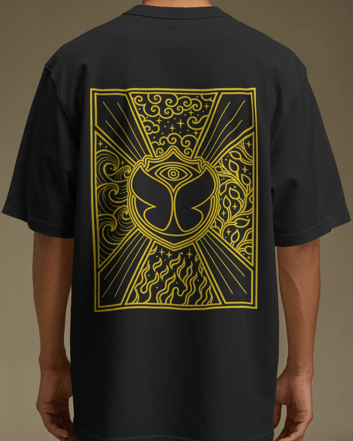 A person wearing a black short-sleeve T-shirt shown from the back, featuring a large yellow graphic print with an eye motif and ornate line art on the back. Nitorious Atelier Tomorrowland Concert collection