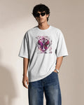 A man wearing a white short-sleeve t-shirt with a pink and purple psychedelic eye graphic on the chest, paired with blue jeans and black sunglasses, standing against a beige background.