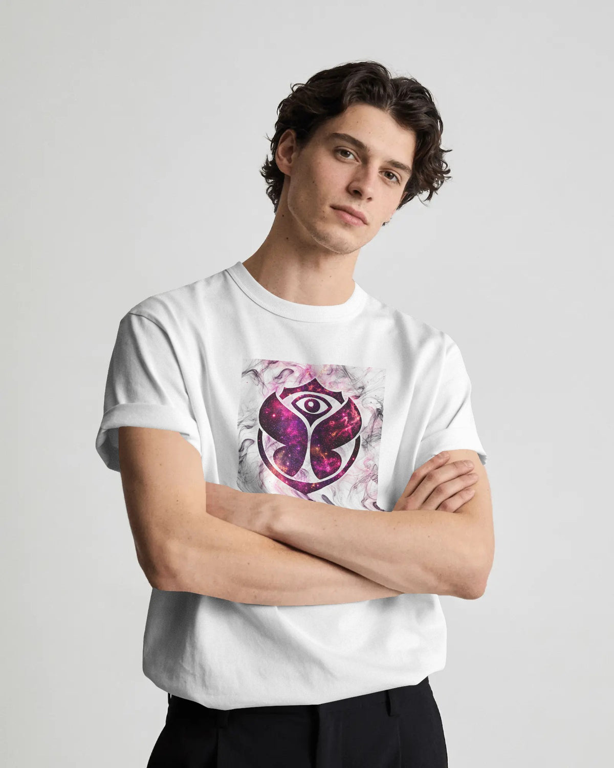 A young man with dark curly hair wearing a white crewneck t-shirt with a purple and pink eye graphic on the front, posed with arms crossed against a light gray background. Tomorrowland Thailand Tshirt