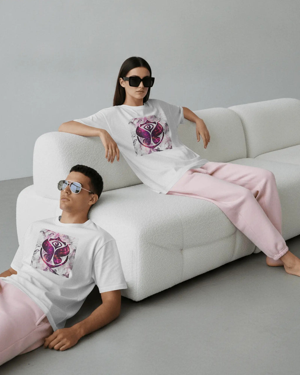 A man and a woman wearing matching white graphic T-shirts and light pink sweatpants, posed on a white textured sofa in a minimalist studio setting. Buy Tomorrowland Thailand Tshirt
