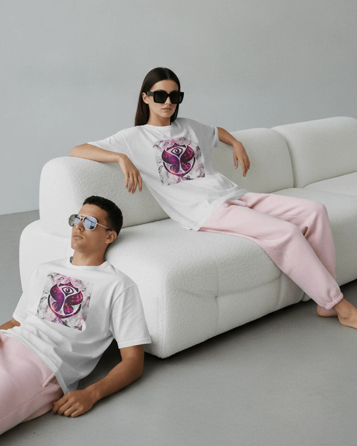 A man and a woman wearing matching white graphic T-shirts and light pink sweatpants, posed on a white textured sofa in a minimalist studio setting. Buy Tomorrowland Thailand Tshirt