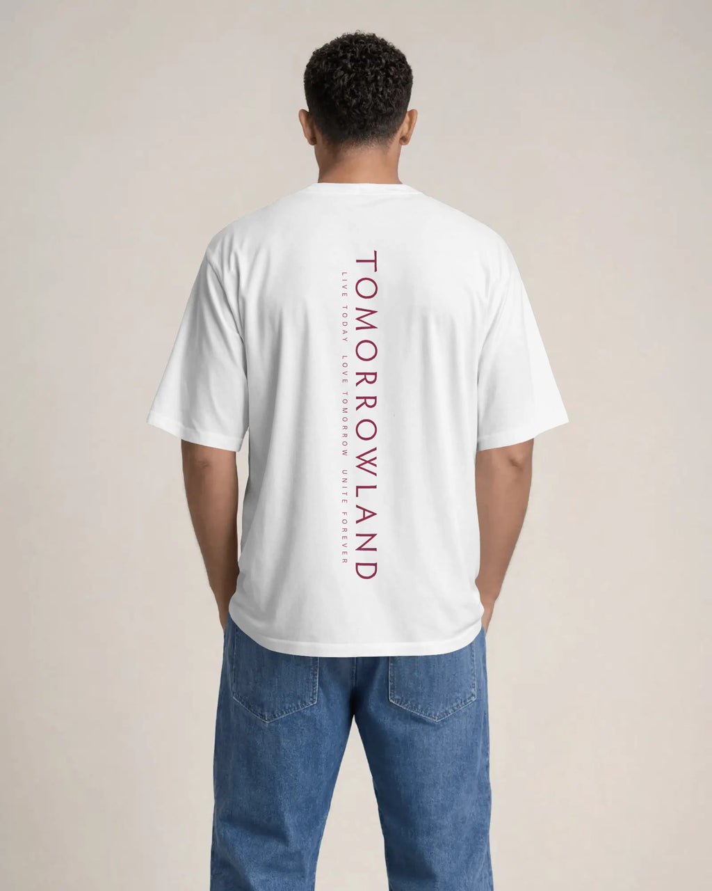 A man viewed from behind wearing a plain white short-sleeve t-shirt and blue jeans, standing against a solid light beige background. Tomorrowland Thailand Tshirt
