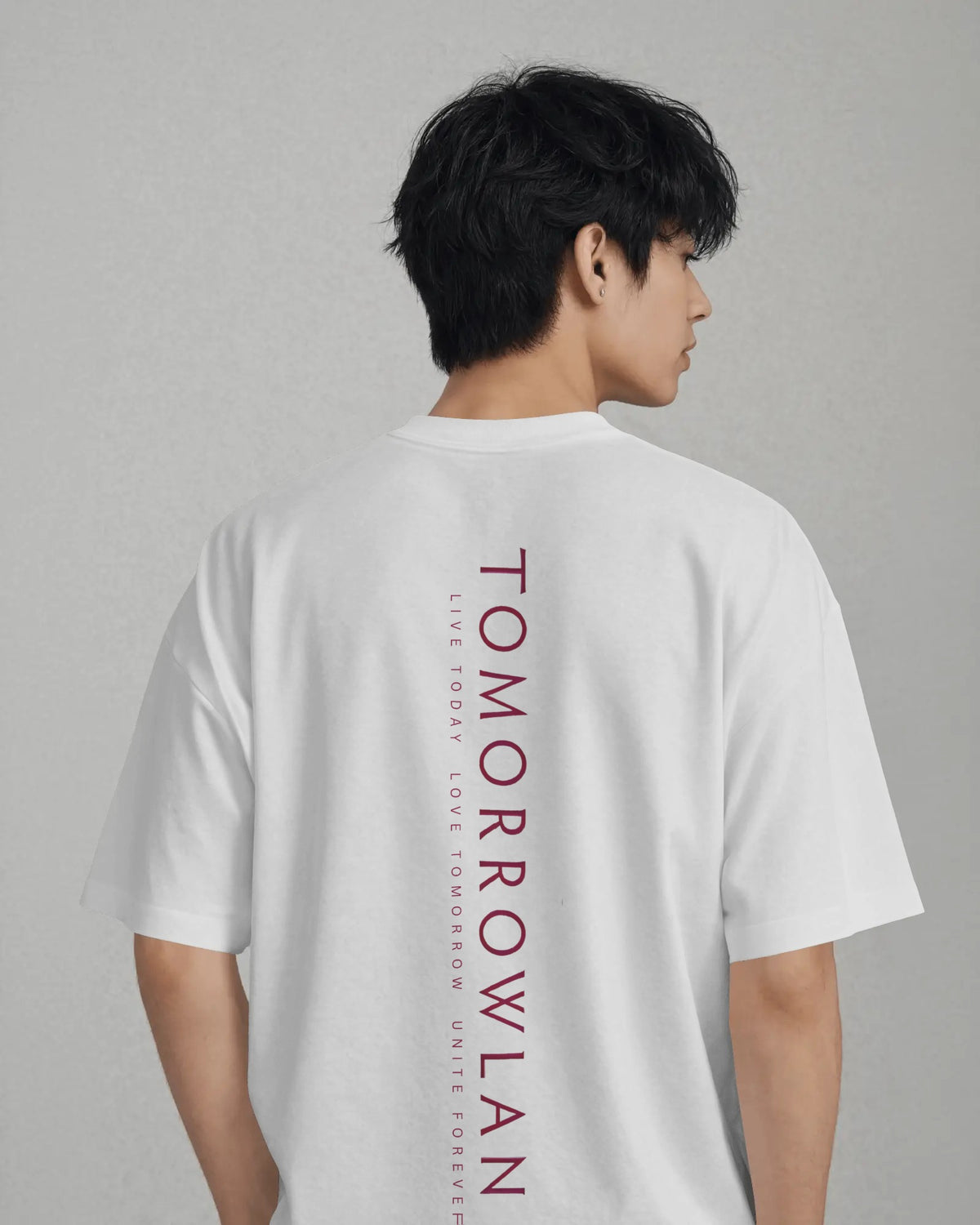 A man wearing a white oversized t-shirt, viewed from the back. The shirt features a vertical graphic print on the back in dark red text reading 'TOMORROWLAND' with smaller text above and below.