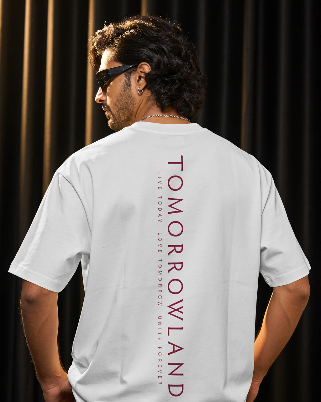A man wearing a white t-shirt with 'TOMORROWLAND' printed vertically in dark red on the back, photographed from behind against a dark curtain background.