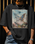A person wearing an oversized black graphic T-shirt with a large rectangular print of a butterfly over a mountain landscape. The shirt is styled with blue jeans, a silver chain necklace, and bracelets, set against a dark background with vertical gold-toned light bars.