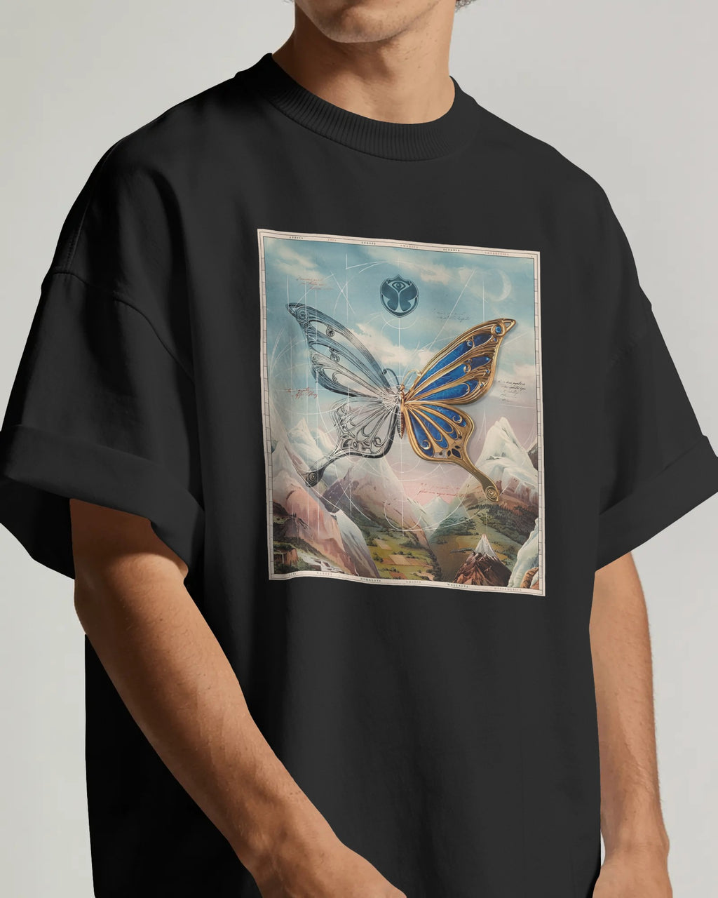 A person wearing an oversized black t-shirt with a large square graphic print on the front. The print features a surreal butterfly over a mountain landscape under a cloudy sky with a moon.