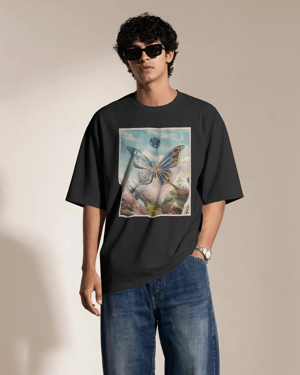 A man wearing a black oversized t-shirt with a colorful butterfly graphic print on the front, paired with blue jeans and black sunglasses, standing against a neutral beige background. Buy Latest Tomorrowland Thailand Tshirt