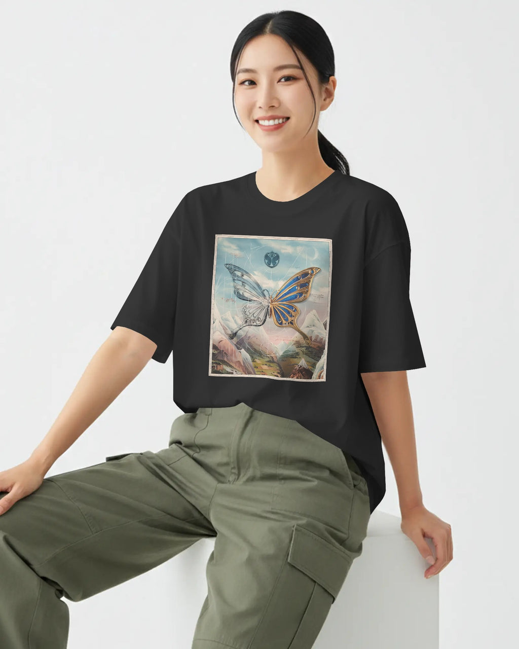 A woman wearing a black oversized t-shirt with a colorful butterfly graphic print on the front, paired with olive green cargo pants, sitting against a plain white background.