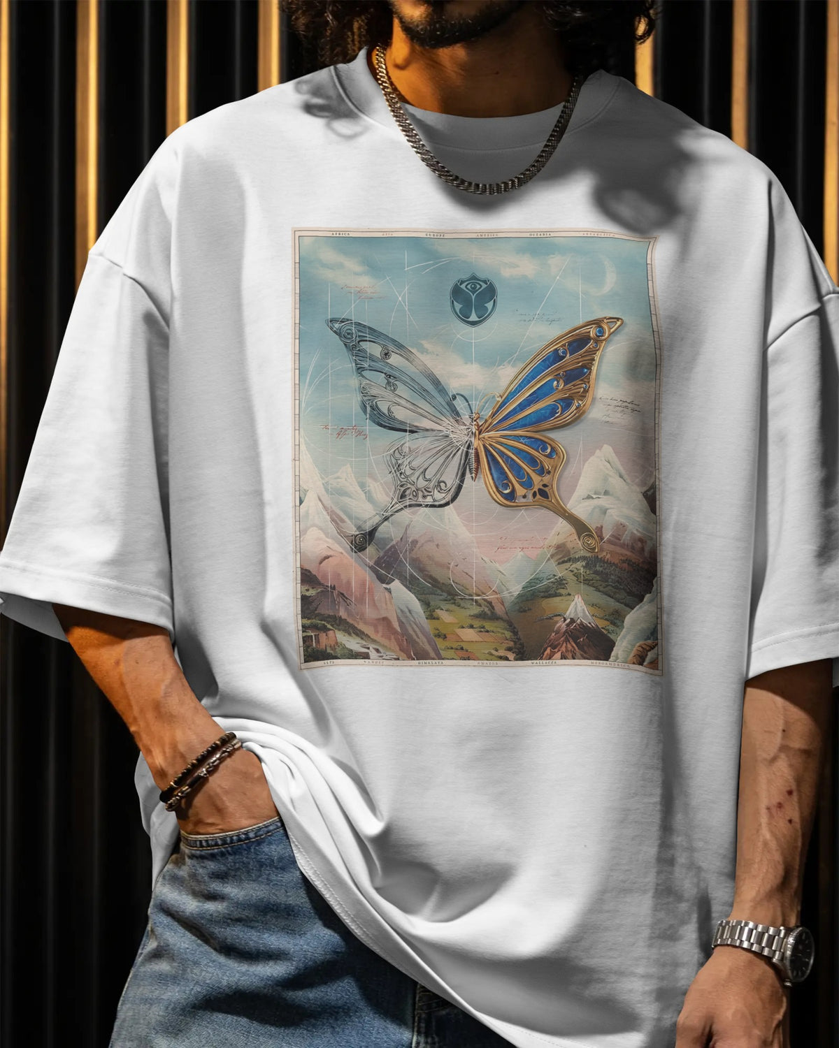 A person wearing an oversized white graphic T-shirt with a large butterfly and mountain print on the front, paired with blue jeans, shown against a dark striped background. Buy Tomorrowland Thailand Tshirt