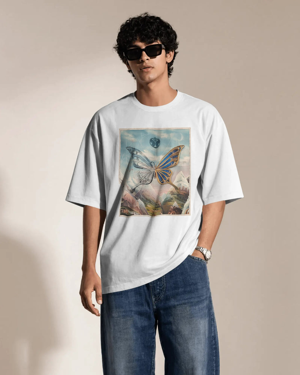 A man modeling an oversized white t-shirt with a colorful butterfly graphic print on the front, paired with blue jeans and black sunglasses, set against a warm beige background.