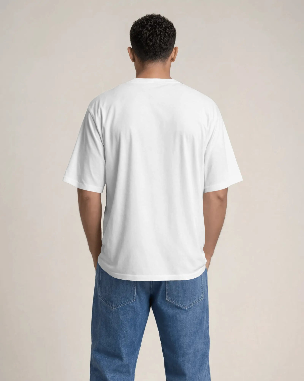 A man viewed from behind wearing a plain white short-sleeve t-shirt and blue jeans, standing against a solid light beige background.