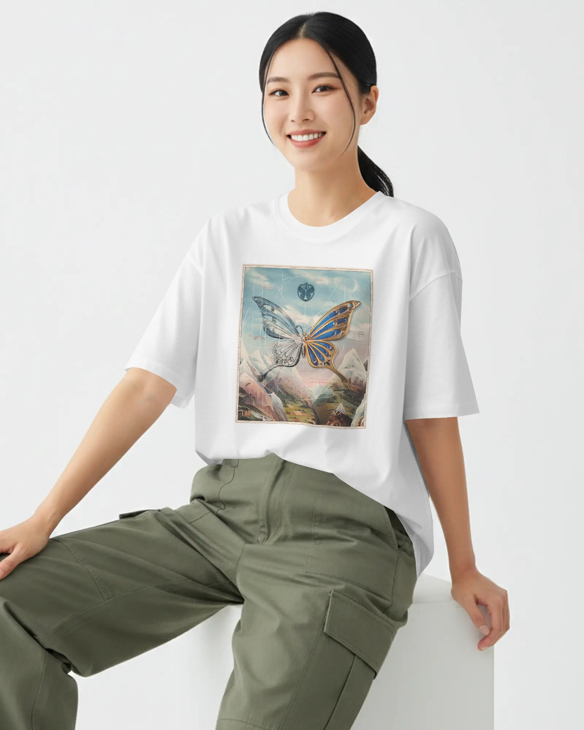 A woman wearing a white short-sleeve graphic T-shirt with a colorful butterfly print on the front, paired with olive green cargo pants, seated against a clean white background.