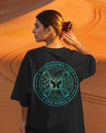 A woman photographed from behind wearing an oversized black T-shirt with a large circular teal graphic on the back that reads “TOMORROWLAND ONE WORLD.” She is standing outdoors against a warm orange sand dune background.