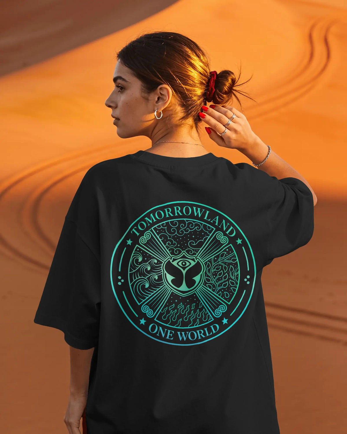 A woman photographed from behind wearing an oversized black T-shirt with a large circular teal graphic on the back that reads “TOMORROWLAND ONE WORLD.” She is standing outdoors against a warm orange sand dune background.