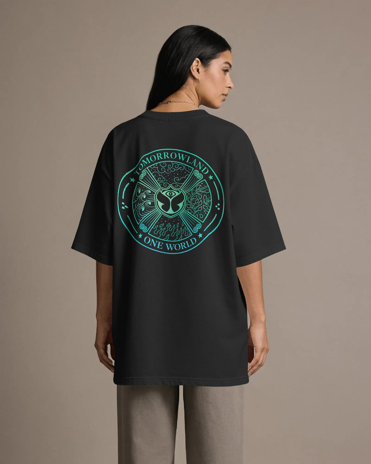 A woman shown from the back wearing an oversized black T-shirt with a large circular teal graphic on the back that reads 'Tomorrowland One World,' paired with light gray pants, against a plain beige studio background.