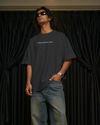 A man wearing an oversized black Tomorrowland t-shirt and blue jeans stands in front of black curtains, with his hand in his pocket.