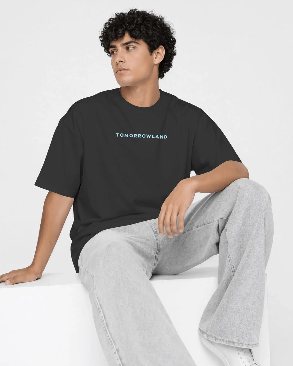 A man wearing a black crewneck t-shirt with a red 'TOMORROWLAND' logo on the chest, paired with light gray jeans, sitting on a white block against a white studio background.