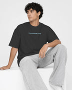 A man wearing a black crewneck t-shirt with a red 'TOMORROWLAND' logo on the chest, paired with light gray jeans, sitting on a white block against a white studio background.