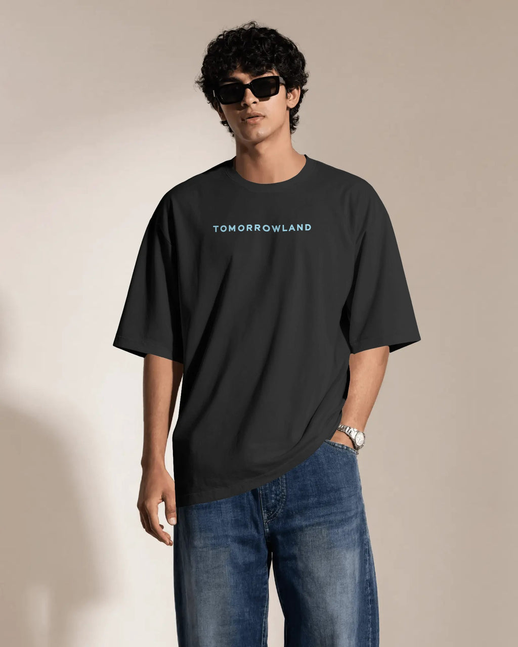 A man wearing a black Tomorrowland t-shirt and blue jeans, standing against a beige studio background with a warm spotlight.
