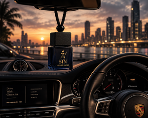 A black car air freshener bottle labeled 'SILENT SIN VELVET SMOKE' hanging from the rearview mirror inside a Porsche car at sunset, with a city skyline reflected in the windshield. Silent Sin Automotive Elixir Car Fragrance