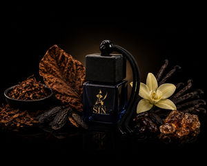 A dark blue glass perfume bottle with a black cap and a black cord, labeled 'Silent SIN' in gold, styled with tobacco leaves, vanilla beans, a vanilla flower, and amber pieces on a black background. Silent Sin Automotive Elixir Car Fragrance