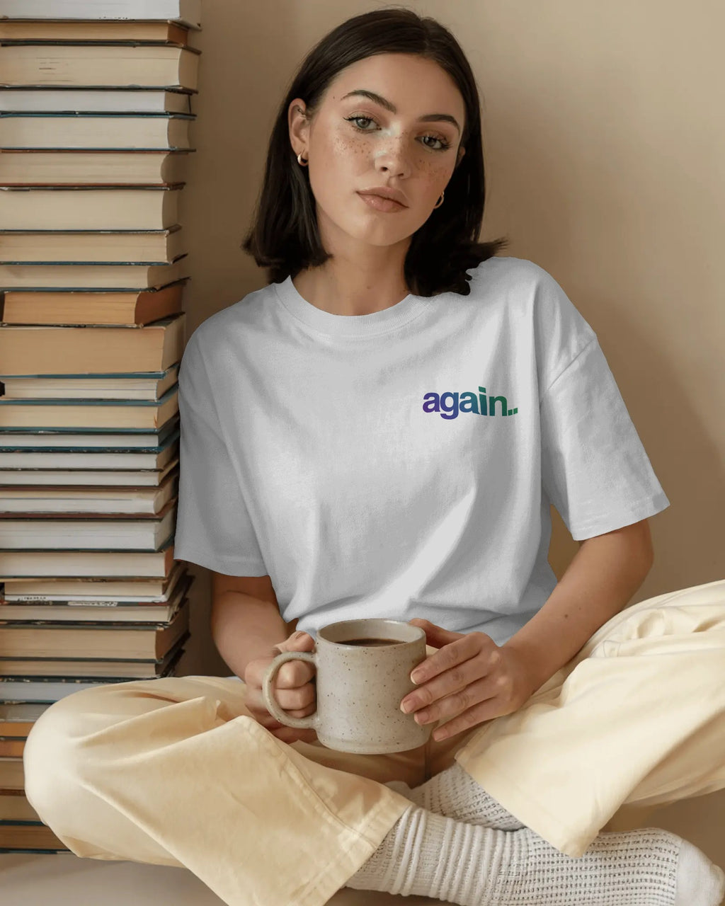 A woman sits cross-legged holding a ceramic mug, wearing a white crewneck t-shirt with the word 'again.' printed in black lowercase letters on the chest, paired with cream-colored pants and knit socks. Fred Again India concert Tshirt  by Nitorious Atelier