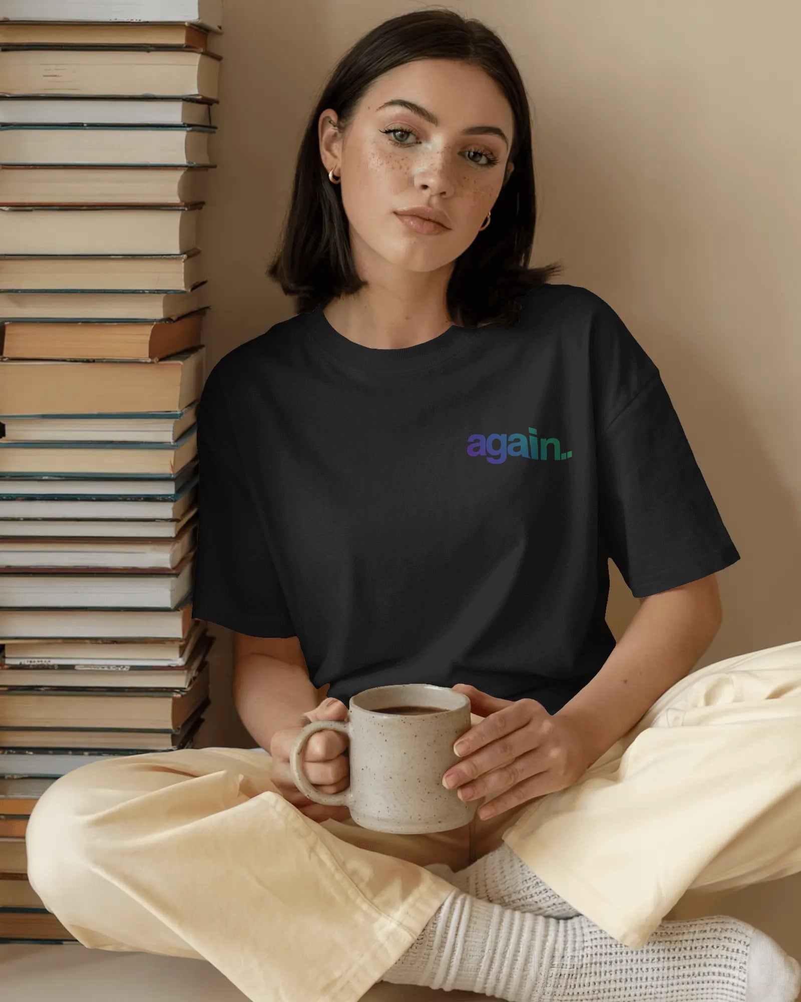 A woman sits cross-legged holding a ceramic mug, wearing a black t-shirt with a small 'again.' logo in purple and green on the chest, paired with cream-colored pants and knit socks. Fred Again India concert Tshirt  by Nitorious Atelier