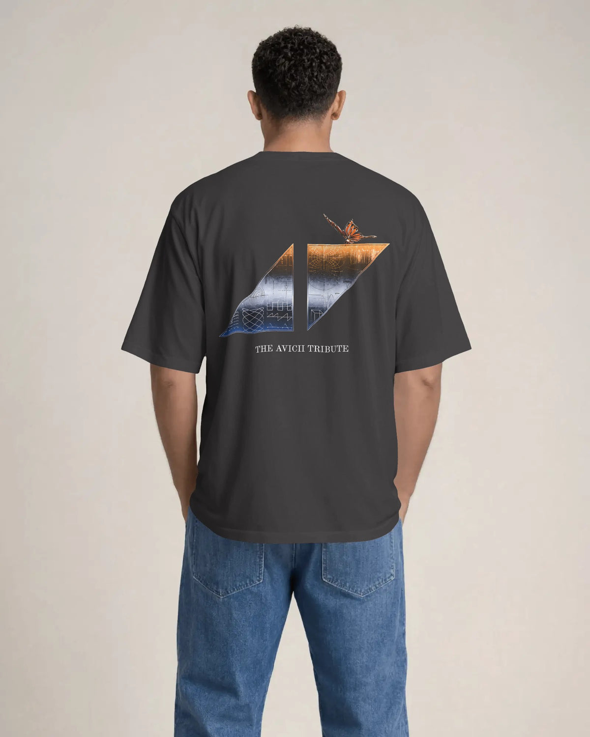 A man viewed from behind wearing a dark charcoal gray t-shirt with a large graphic on the back and blue jeans, standing against a plain beige studio background. The Avicii Tribute Tomorrowland Tshirt.